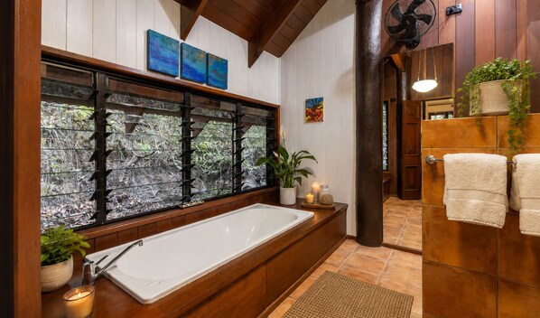 Un baño moderno con una bañera grande, detalles de madera, un cerramiento de vidrio con plantas y un ventilador.