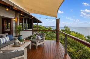 A wooden deck with outdoor furniture, a pergola with string lights, and a view of the sea.