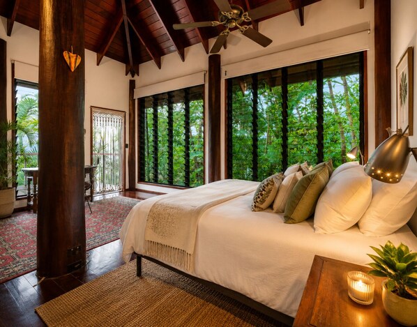 A bedroom with a bed, a ceiling fan, large windows, a wooden floor, and a view of greenery outside.