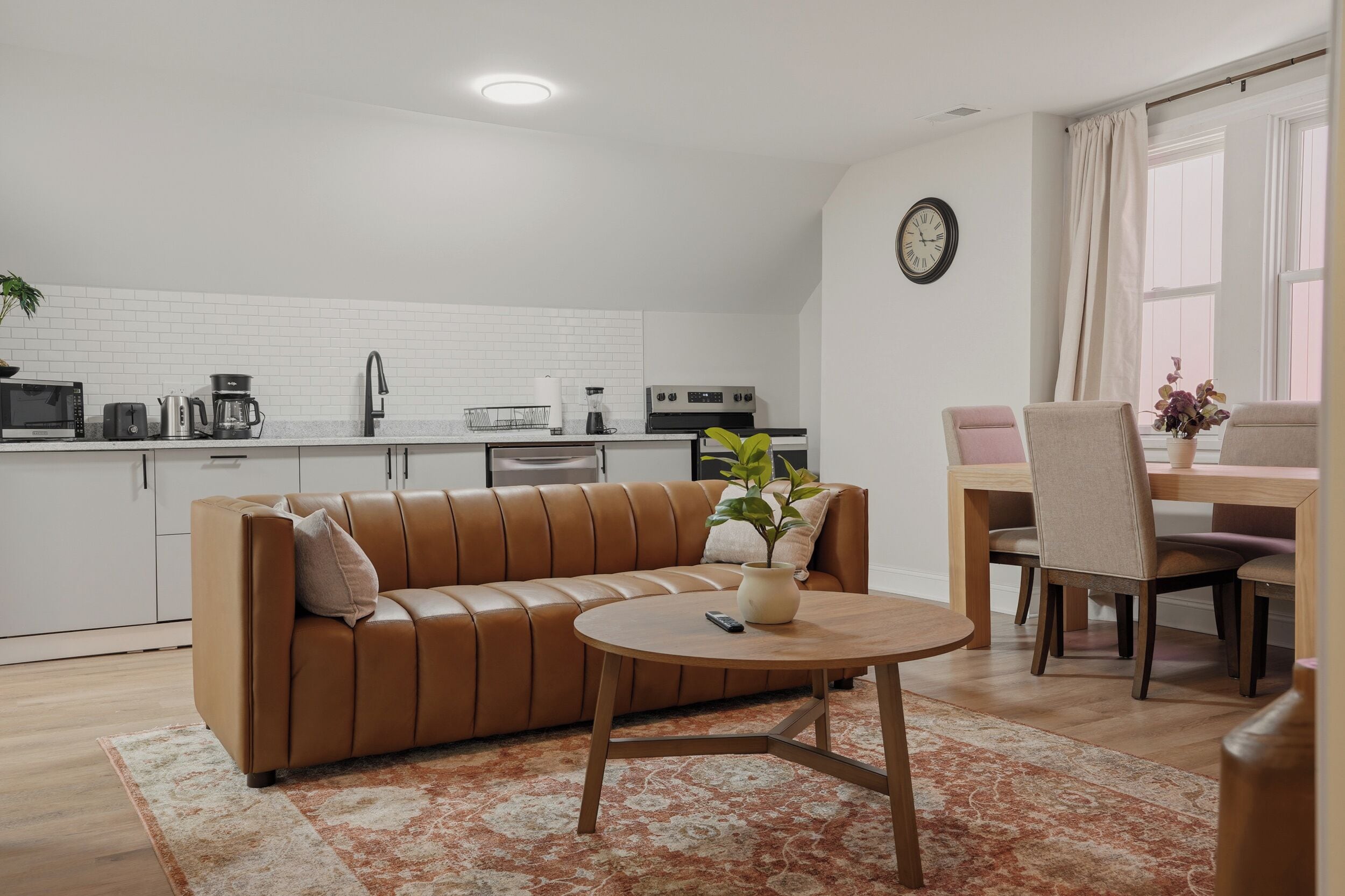 A modern living space with a brown leather sofa, a round wooden coffee table, and a kitchen area with white cabinetry and a brick backsplash.