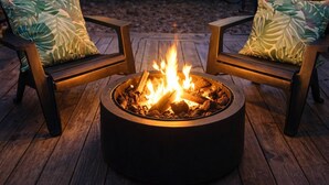 Dos sillas Adirondack con cojines de estampado de hojas, un hogar circular con llamas y una terraza de madera.