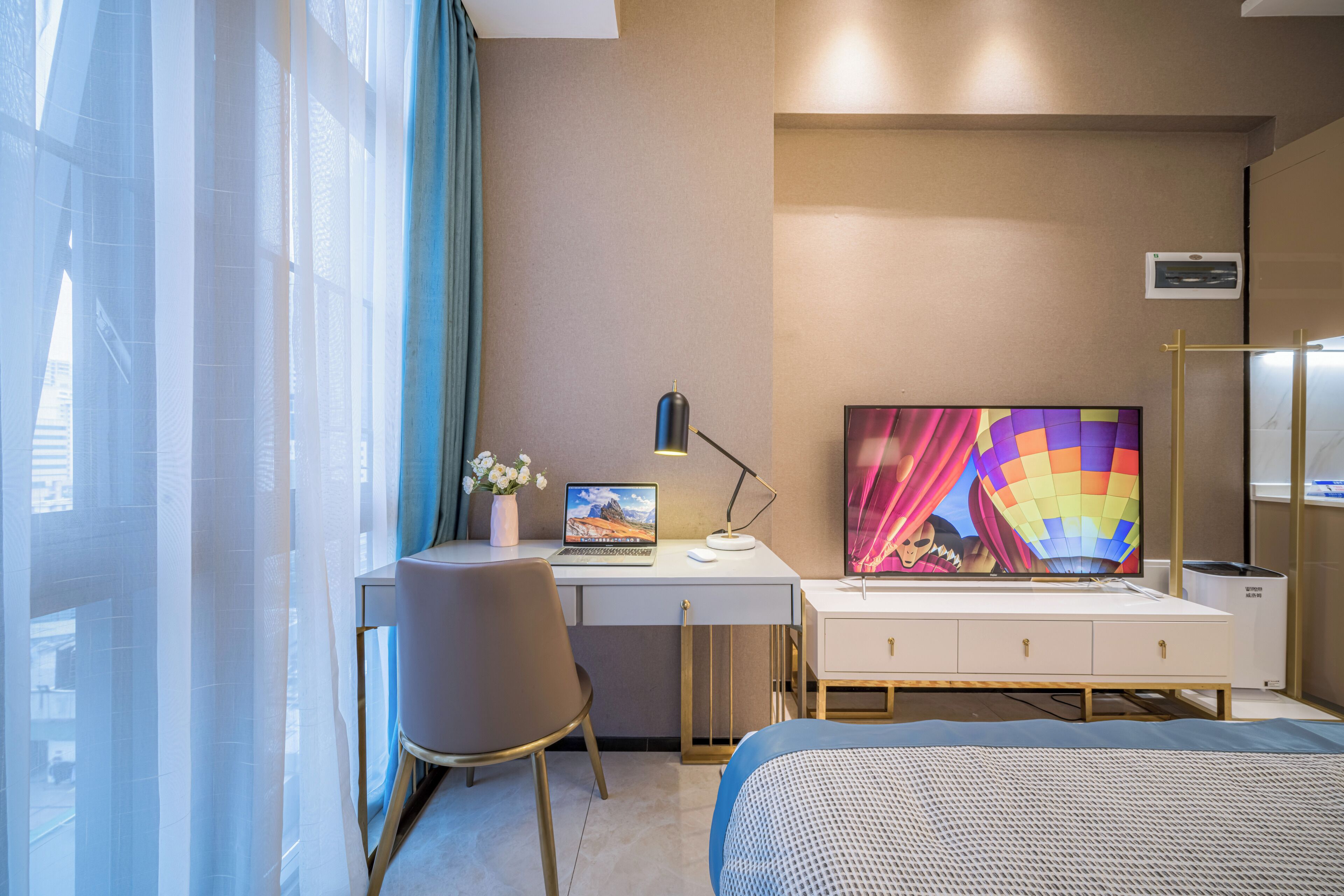 A hotel room with a bed, a desk with a laptop, a chair, a TV, and a window with curtains.