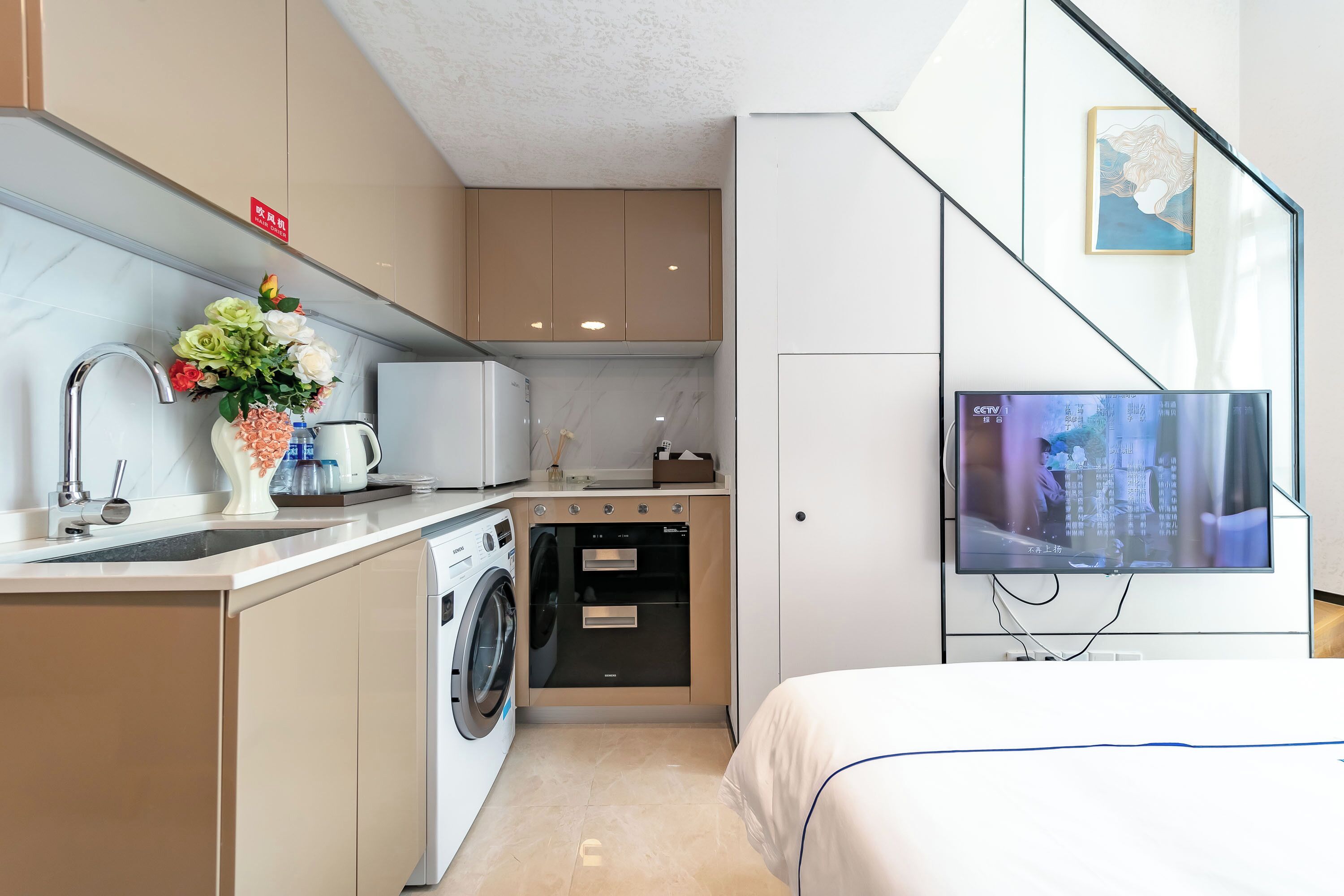 A compact hotel room with a kitchenette, a washing machine, a flat-screen TV, and a bed.