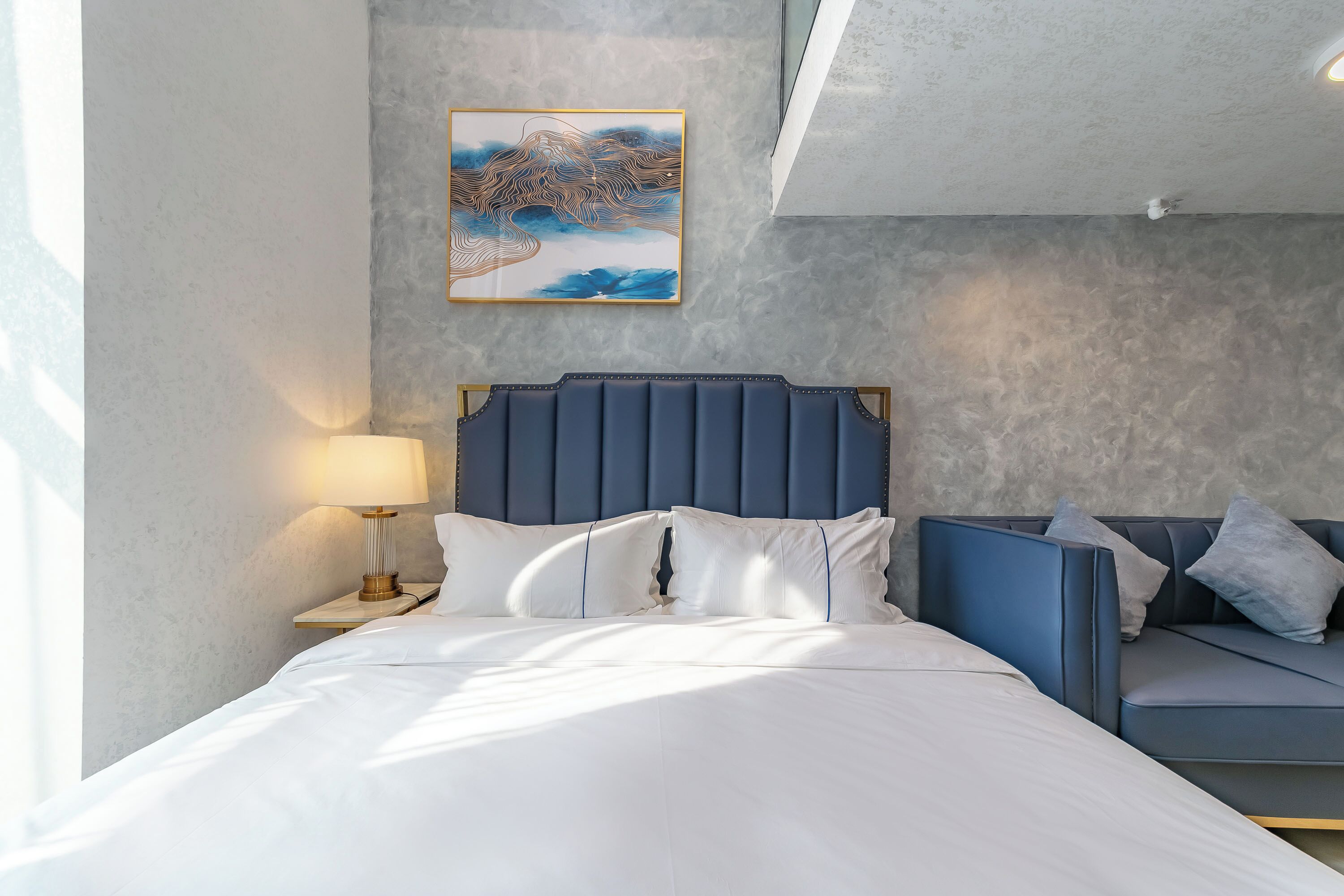 A bedroom with a bed, a nightstand with a lamp, a blue headboard, a sofa, and a wall-mounted artwork.