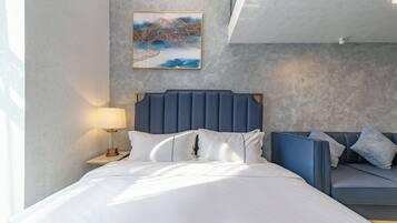 A bedroom with a bed, a nightstand with a lamp, a blue headboard, a sofa, and a wall-mounted artwork.