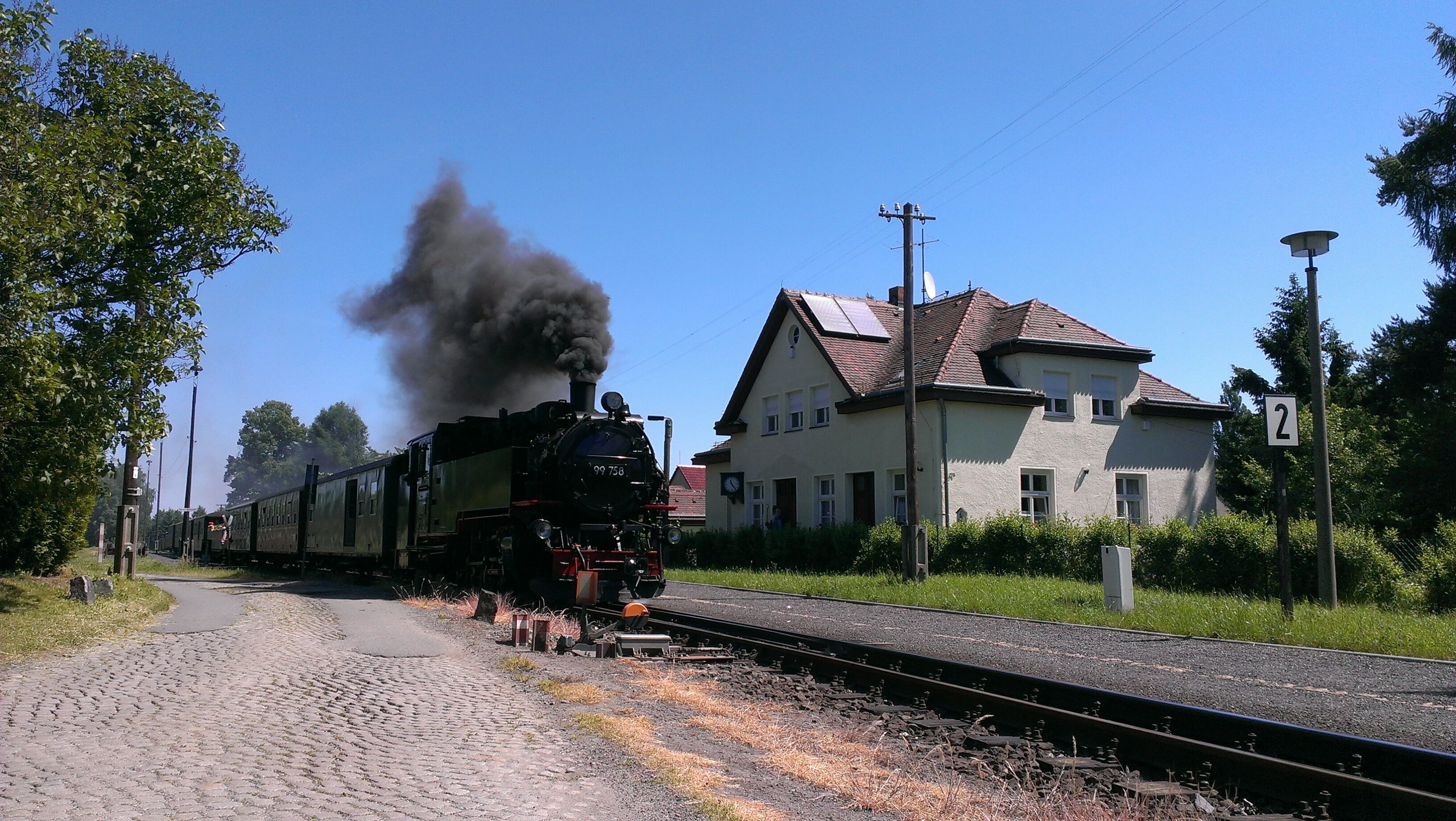 A steam locomotive with the number 99 758 on a railway track, a house with a number 2 sign, and a clear blue sky.