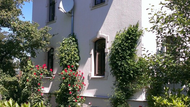 A two-story building with a red roof, multiple windows, and a garden with various plants and flowers.