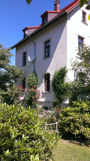 A two-story building with a red roof, multiple windows, and a garden with various plants and flowers. - Vacation apartment for 2-4 persons (40 m²) in Olbersdorf/Zittauer Gebirge (Olbersdorf)