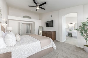 A bedroom with a bed, a ceiling fan, a TV, a dresser, and a bathroom visible through an archway.