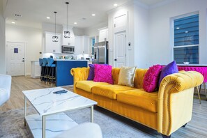 A modern living room with a yellow sofa, a white coffee table, and a kitchen in the background. - 4BR Houston Home - Hot Tub, Outdoor Movies & Karaoke Near NRG/TMC (Houston)