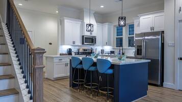 A modern kitchen with white cabinets, a central island with blue bar stools, stainless steel appliances, and a wooden staircase.