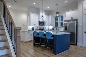 A modern kitchen with white cabinets, a central island with blue bar stools, stainless steel appliances, and a wooden staircase. - 4BR Houston Home - Hot Tub, Outdoor Movies & Karaoke Near NRG/TMC (Houston)