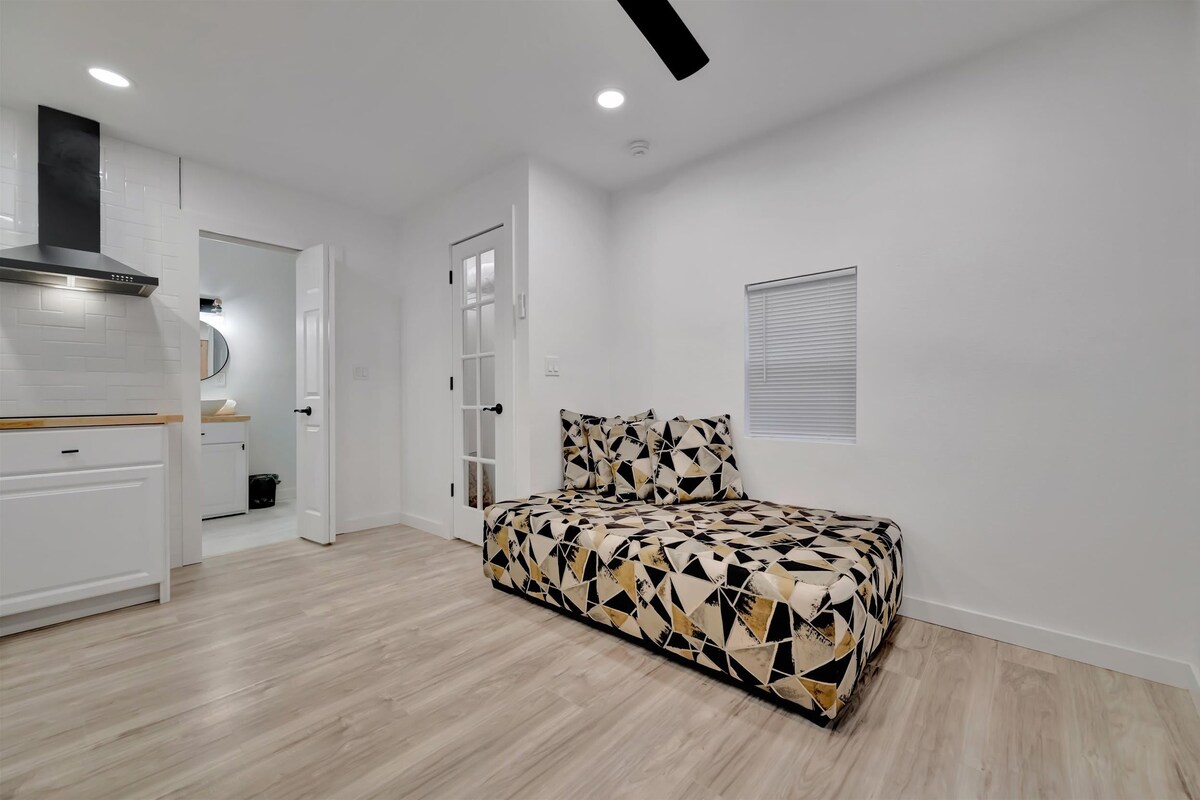 A modern bedroom with a geometric-patterned bed, a kitchenette with white cabinets, and a ceiling fan.