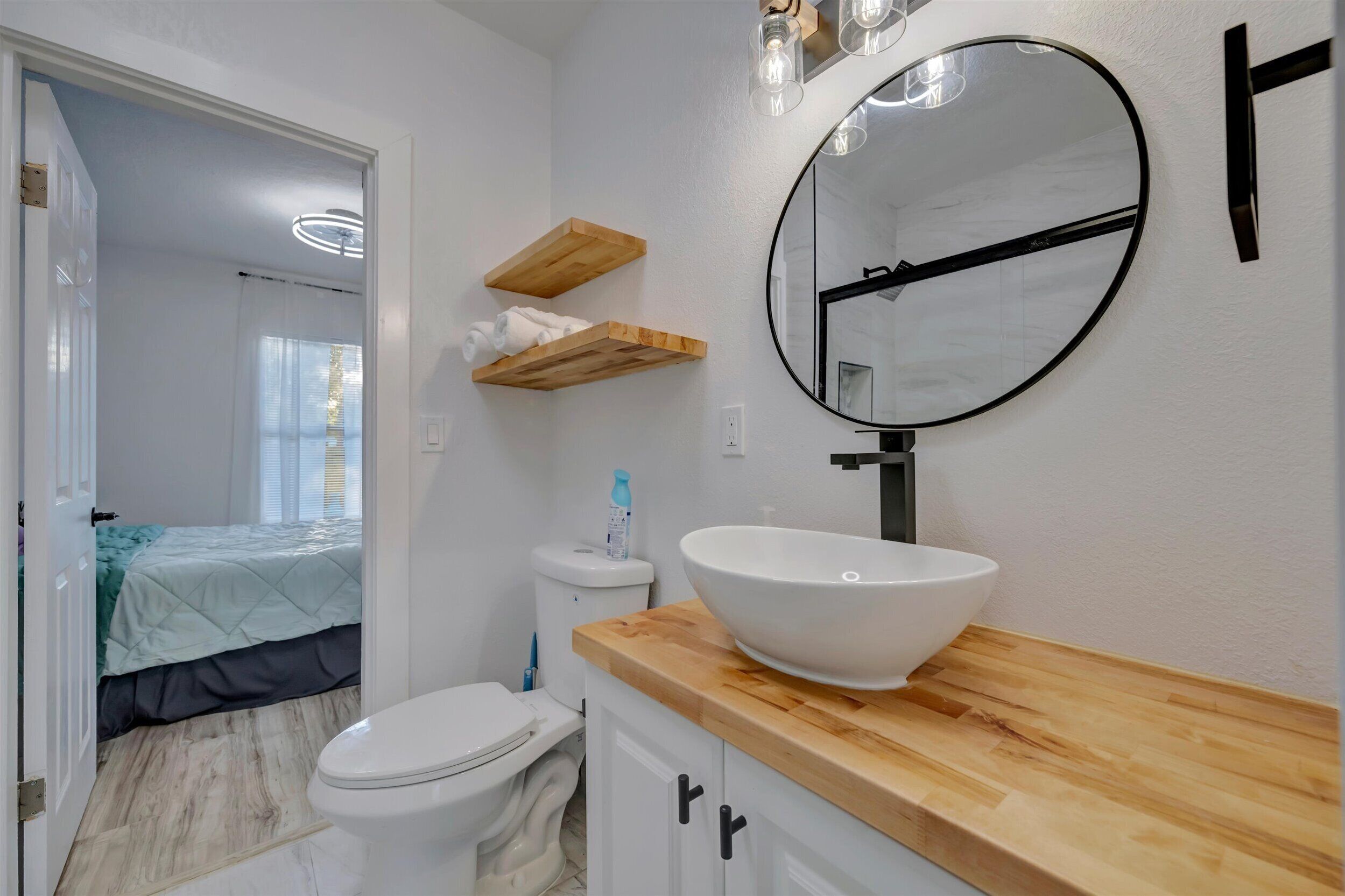 A bathroom with a white sink, wooden shelves, a round mirror, and a toilet.