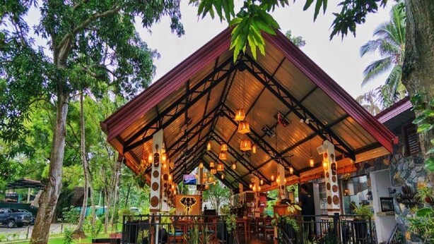 A covered outdoor seating area with tables and chairs, surrounded by greenery.