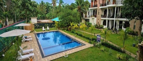 A swimming pool with lounge chairs, surrounded by lush greenery and a multi-story building with balconies.
