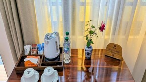 A hotel room with a wooden table set with two white teacups, a kettle, a water bottle, a vase with a flower, and a tray with coffee packets and a non-smoking sign.