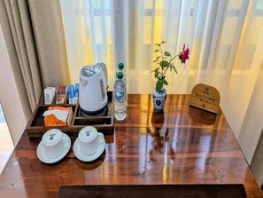 A hotel room with a wooden table set with two white teacups, a kettle, a water bottle, a vase with a flower, and a tray with coffee packets and a non-smoking sign. - The Grand Toscana - Udawalawa (Udawalawa)