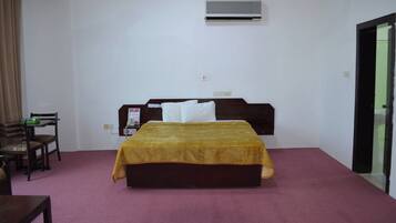 A hotel room with a bed, a desk, a chair, an air conditioning unit, and a door.
