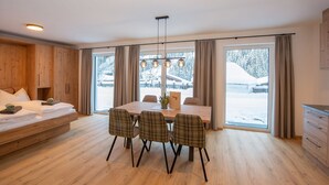 Ein modernes Zimmer mit einem Holzbett, einem Esstisch mit karierten Stühlen und großen Fenstern mit Vorhängen.