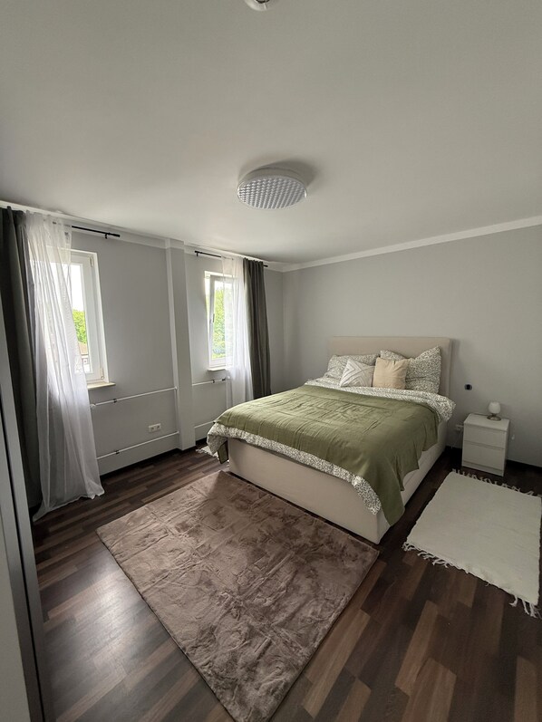 Une chambre à coucher avec un lit, une table de chevet, une fenêtre avec des rideaux et un luminaire au plafond.