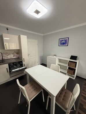 Une cuisine compacte équipée d’un lave-vaisselle, d’un four et de meubles blancs. On y trouve une table à manger avec des chaises, ainsi qu’une petite étagère blanche avec des paniers.