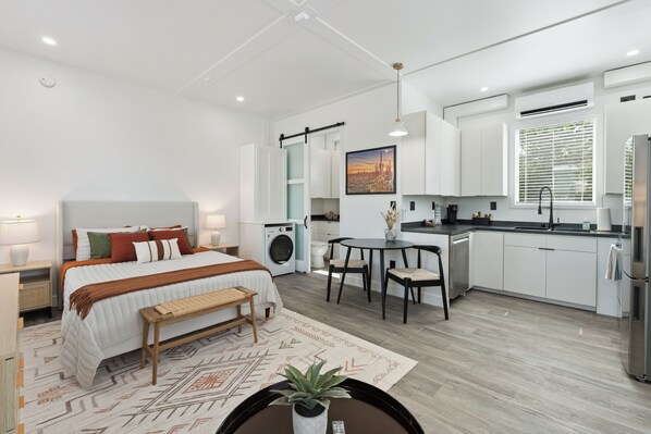 A modern hotel room with a bed, bedside table, bench, and a small dining area with chairs.
