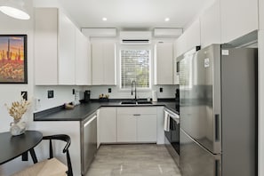 A modern kitchen with white cabinets, a stainless steel refrigerator, and a window with blinds.