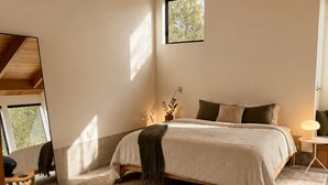 Ein Schlafzimmer mit einem Bett, einem kleinen Tisch, einem Sessel, einem Nachttisch, einem Spiegel und einem Fenster.