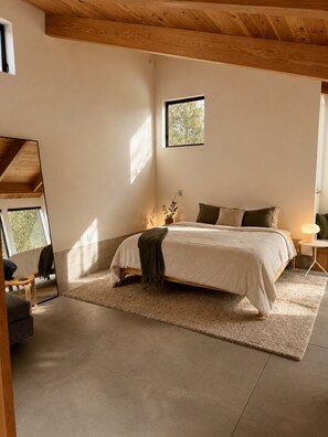 A bedroom with a bed, a small table, a chair, a nightstand, a mirror, and a window.
