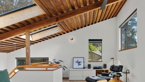 Ein modernes Interieur mit schrägem Dach, Holzbalken, einem Schreibtisch mit Stuhl, einer Couch und einem Fenster mit Blick auf Bäume.