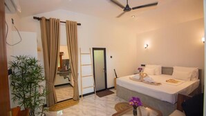A hotel room with a bed, a ceiling fan, a small table with a vase, and a potted plant.