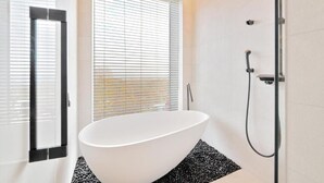 Ein modernes Badezimmer mit einer freistehenden Badewanne, einer Duschkabine aus Glas und einem großen Fenster mit Jalousien.