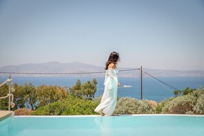 Una persona con un vestido blanco de pie junto a una piscina, con vistas al mar y a las montañas.