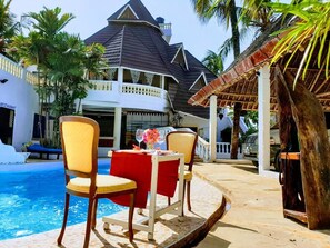 A poolside area with chairs and a table, a building with a dark roof, and palm trees. - Tranquil 1-bedroom cottage with AC in Diani Beach (Diani Beach)