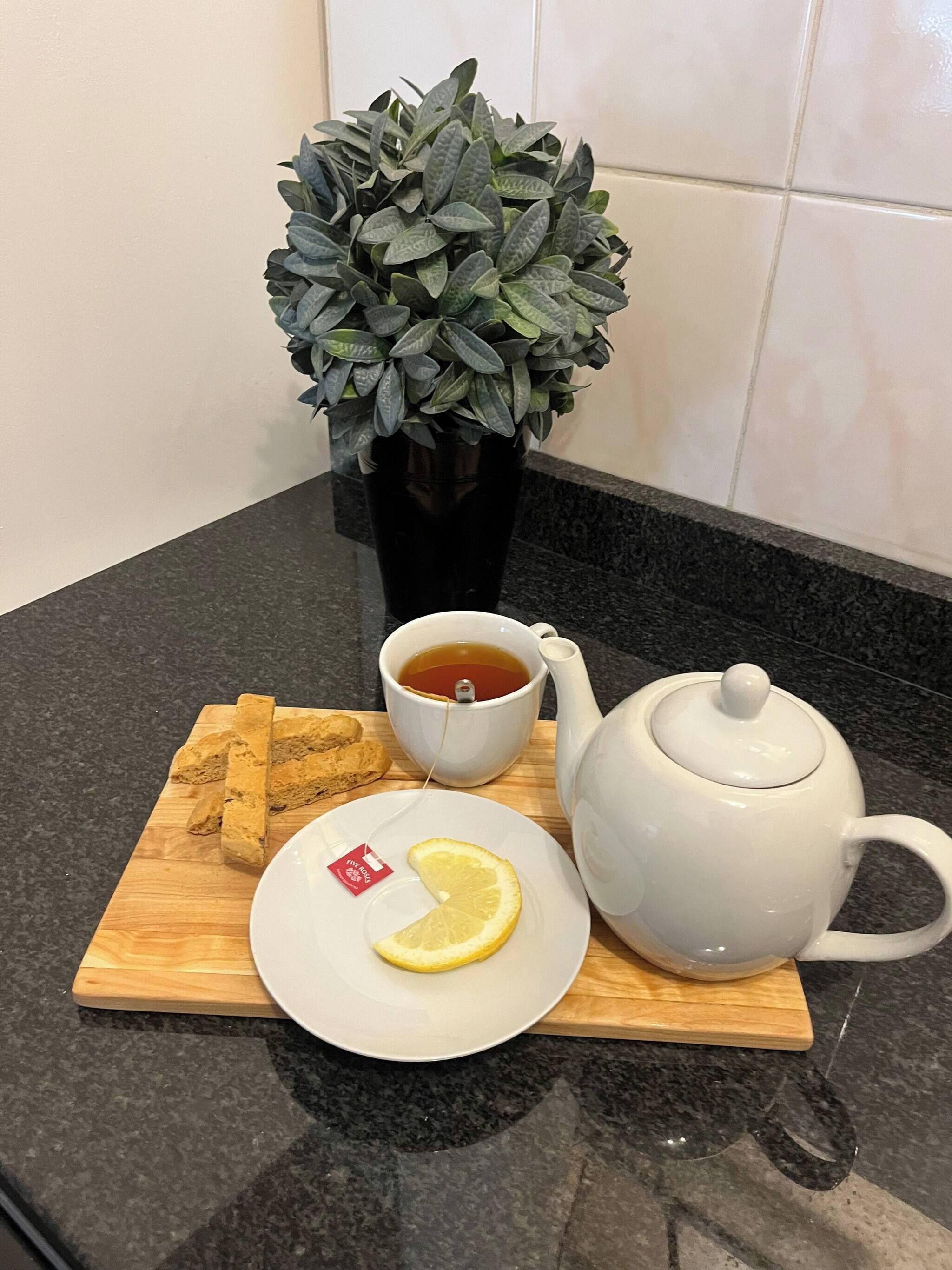 A tray with tea, lemon slice, and biscuits on a wooden board, a white teapot, and a potted plant on a kitchen counter.