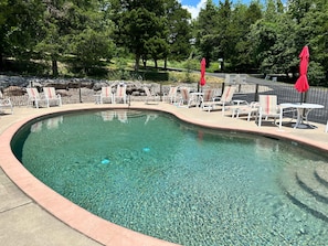 A clear swimming pool with lounge chairs and red umbrellas, surrounded by a stone wall and trees.
