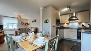Una cocina moderna con armarios blancos, un comedor con mesa de madera dispuesta para cuatro personas, y una sala de estar acogedora con sofá marrón.