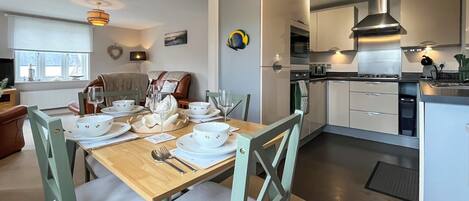 A modern kitchen with white cabinets, a dining area with a wooden table set for four, and a cozy living room with a brown sofa.