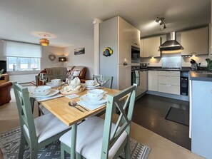 Une cuisine moderne avec des meubles blancs, un coin repas avec une table en bois dressée pour quatre personnes, et un salon confortable avec un canapé marron.