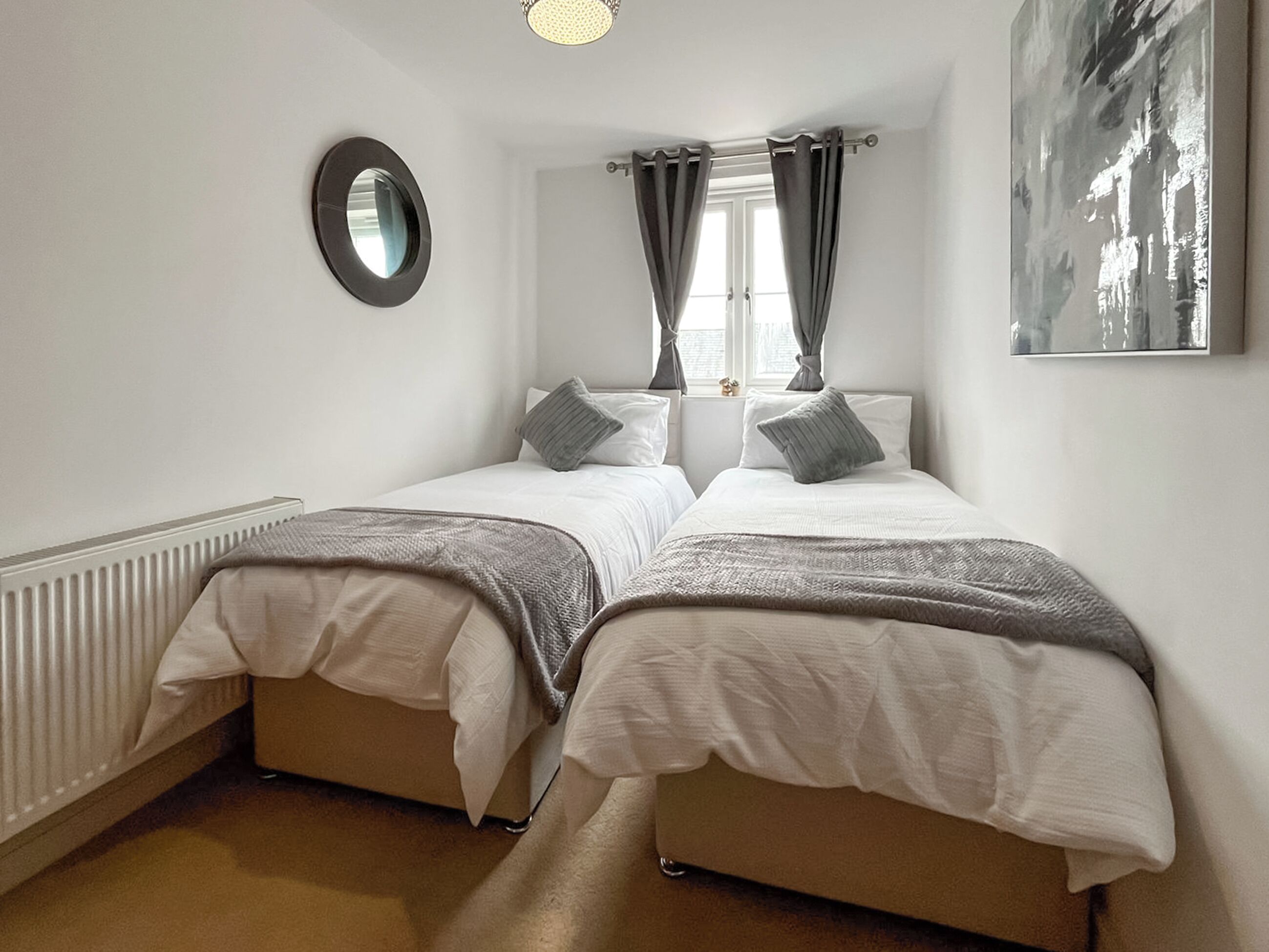 A bedroom with two beds, a round mirror, a window with curtains, and a painting on the wall.