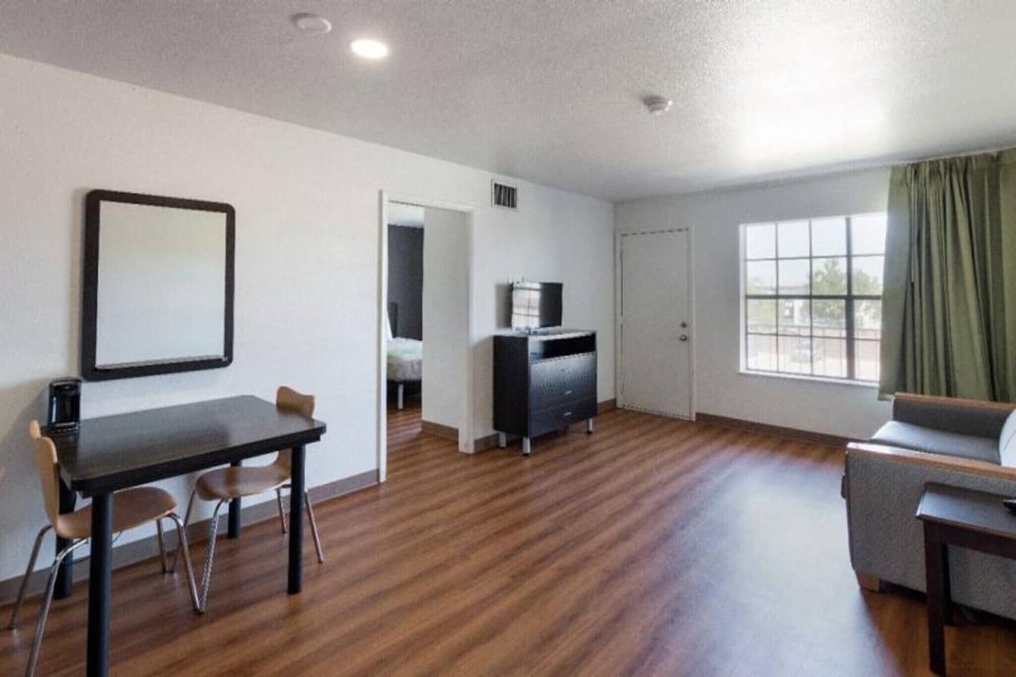 A room with a wooden floor, a dining table with chairs, a sofa, a TV stand, and a window with curtains.