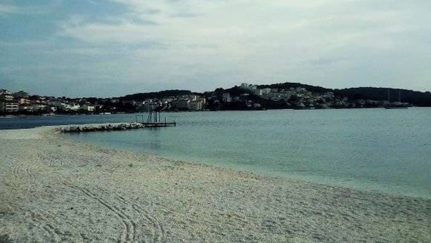 Ein Strand mit klarem Wasser, einer Mole und einer entfernten Hügelkette mit Gebäuden.