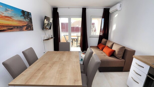 Ein Essbereich mit Holztisch und Stühlen, einer Couch mit Kissen, einer Küchenzeile und einem großen Fenster mit Vorhängen.