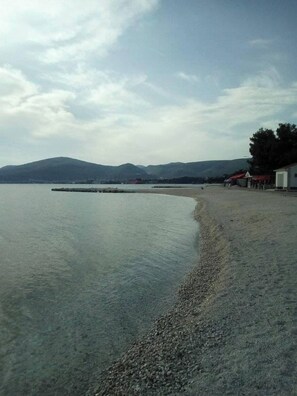 Ein Strand mit Kiesufer, ein Steg, der sich ins ruhige Wasser erstreckt, und in der Ferne Berge unter einem bewölkten Himmel.