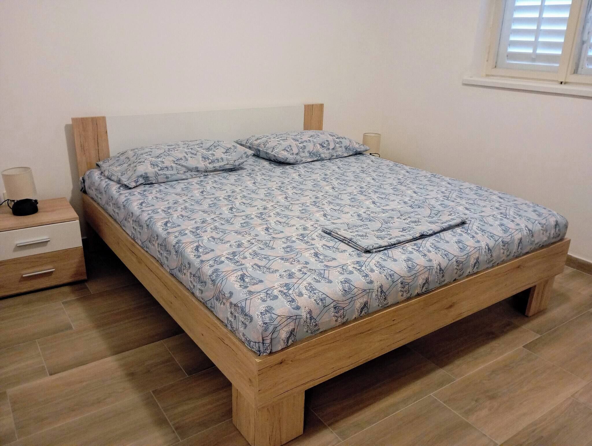 Una cama individual con colcha estampada, cabecera de madera, mesita de noche con lámpara y ventana con persianas.