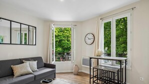 Un salon moderne avec un canapé gris, une horloge et de grandes fenêtres donnant sur un paysage verdoyant.