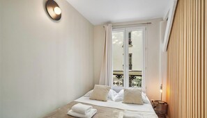Ein Hotelzimmer mit Bett, Fenster mit Vorhängen, Balkon mit Geländer und einer Wandleuchte.
