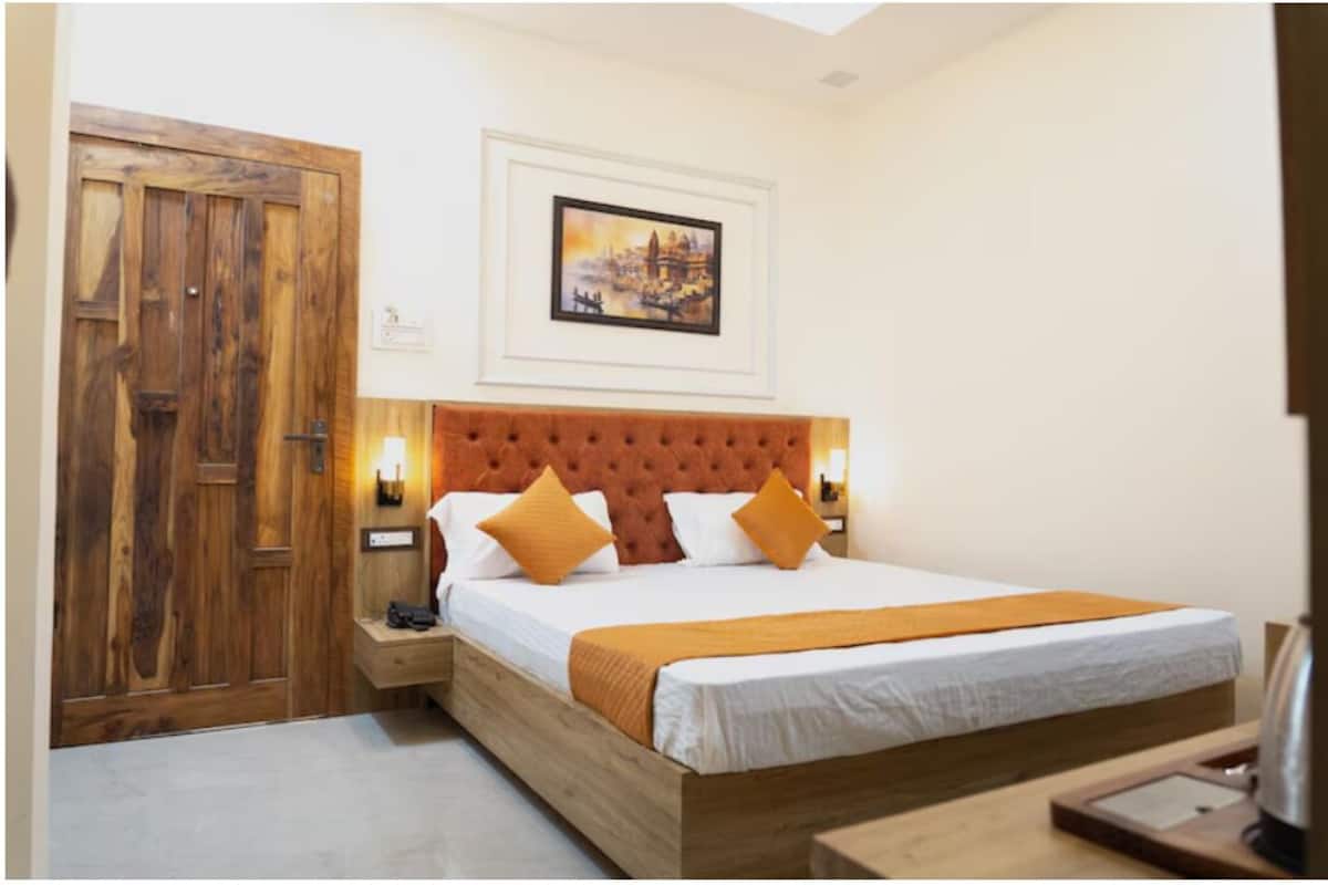 A bedroom with a wooden door, a bed with a tufted headboard, a framed picture on the wall, and a nightstand with a lamp.