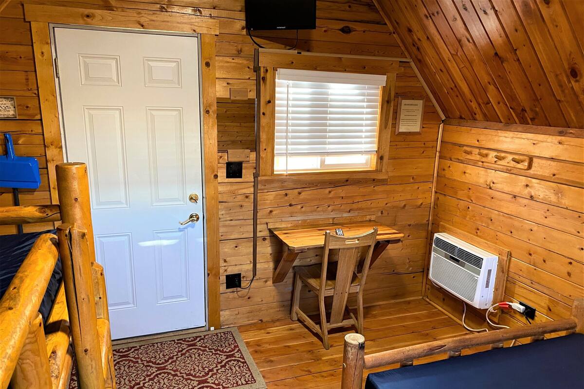 Una cabaña con interior de madera, una puerta, una ventana con persianas, una mesita, una silla, un calentador y una cama.